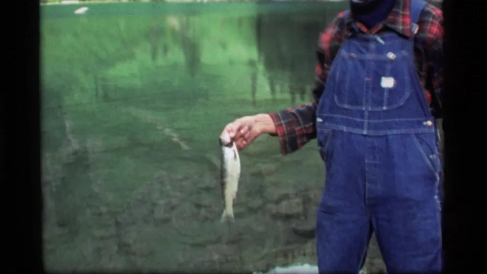 In the 1977 Montana home movie, an adult wearing blue overalls and a plaid shirt holds a small freshly caught fish toward the camera.