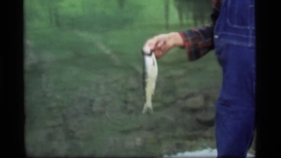 The final 1977 Montana frame crops close on the angler's hand holding the fish beside blue denim overalls and soft green scenery.