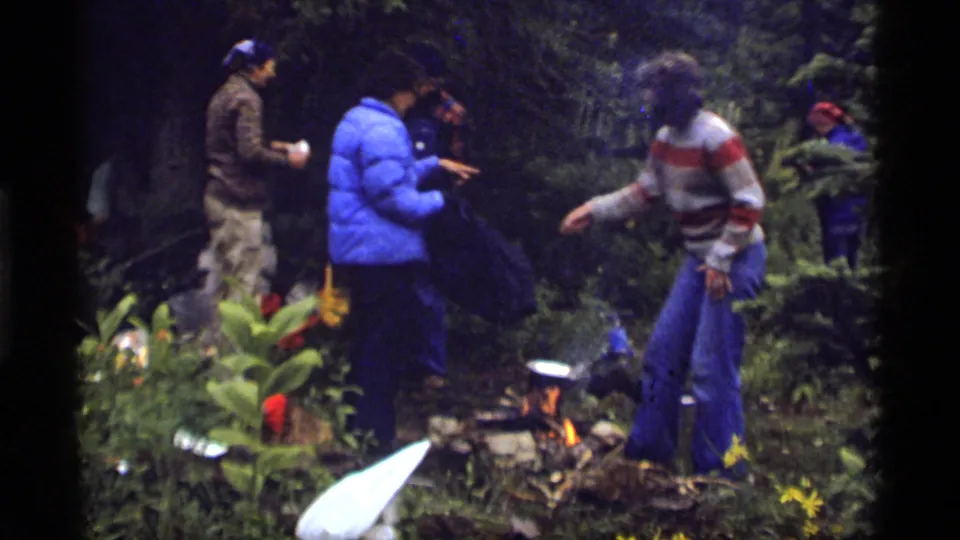 Sapphire Lake campsite frame showing adults near a small fire, with one person in a blue jacket and another in a striped sweater.