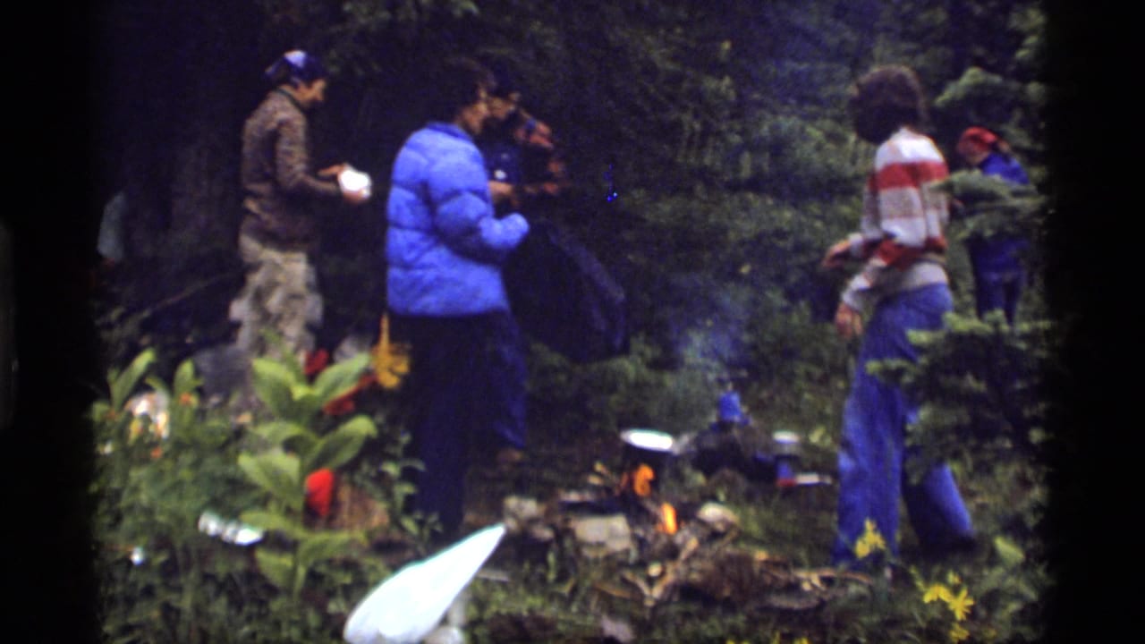 1977 Sapphire Lake camping scene with several people gathered near a small fire and cooking gear in forest underbrush.