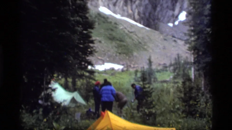 Campers bend and stand near tents in the Sapphire Lake valley, with trees, grass, rocks, and snow patches behind them.