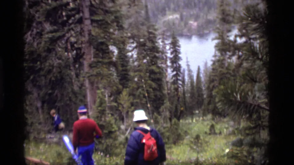 1977 Sapphire Lake hiking footage shows people moving through forest, with a white hat, red backpack, and blue carried object visible.