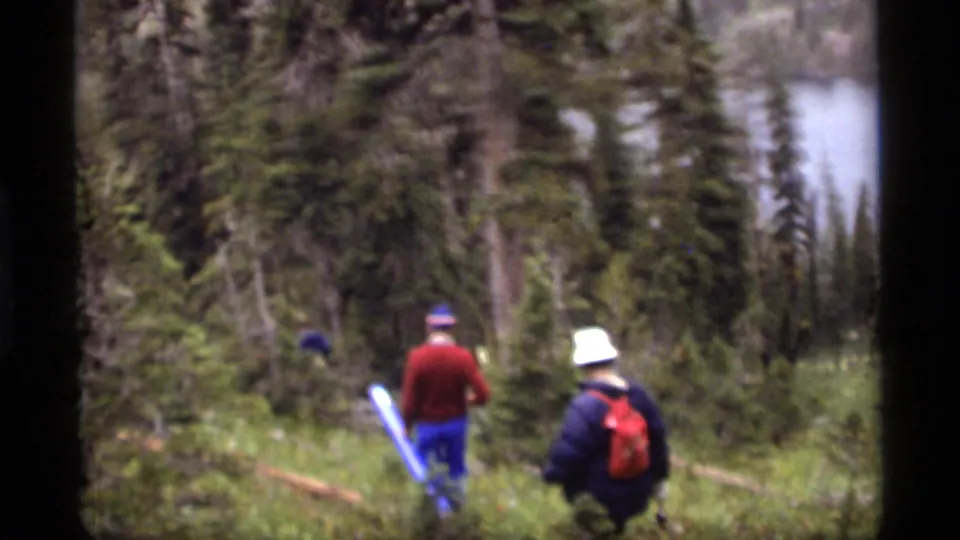 A 1977 8mm frame from Sapphire Lake, Montana, shows hikers in 1970s outdoor clothing walking through wooded terrain.