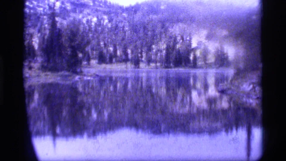 1977 8mm view of Sapphire Lake, Montana, showing a forested shoreline reflected in calm, purple-tinted water.