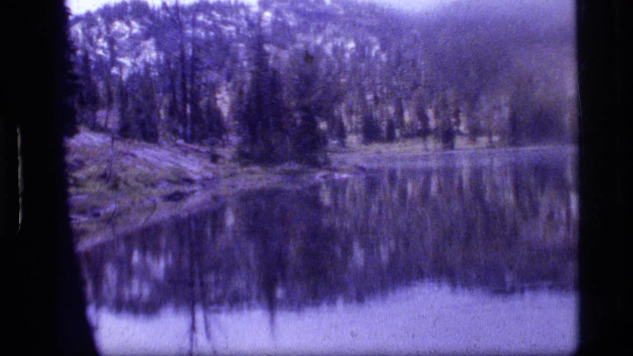 A 1977 8mm view of Sapphire Lake with dense trees and a rocky shore mirrored in the water.