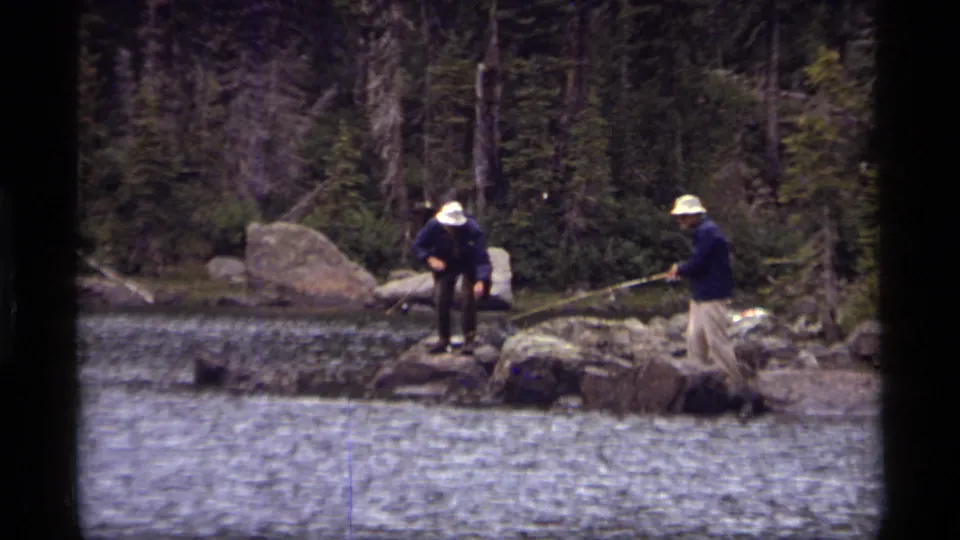 Two adult men wearing hats and dark coats stand on boulders with fishing rods at Sapphire Lake, Montana, in 1977.