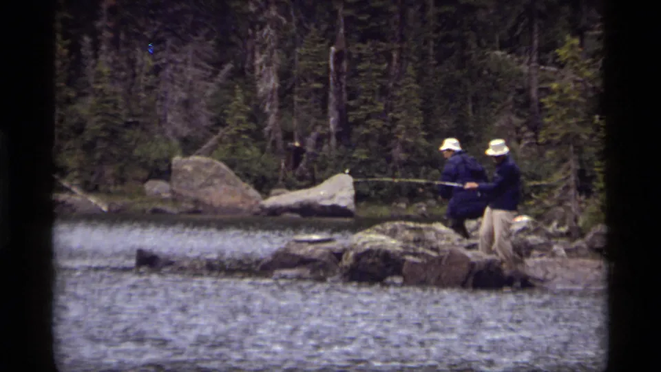 The 1977 home-movie frame shows two adult men fishing from boulders beside Sapphire Lake with trees behind them.