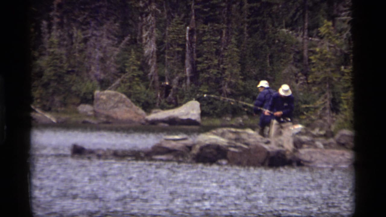 Two people fish from boulders along Sapphire Lake in Montana, with evergreen forest and rippled water visible in 1977 8mm footage.