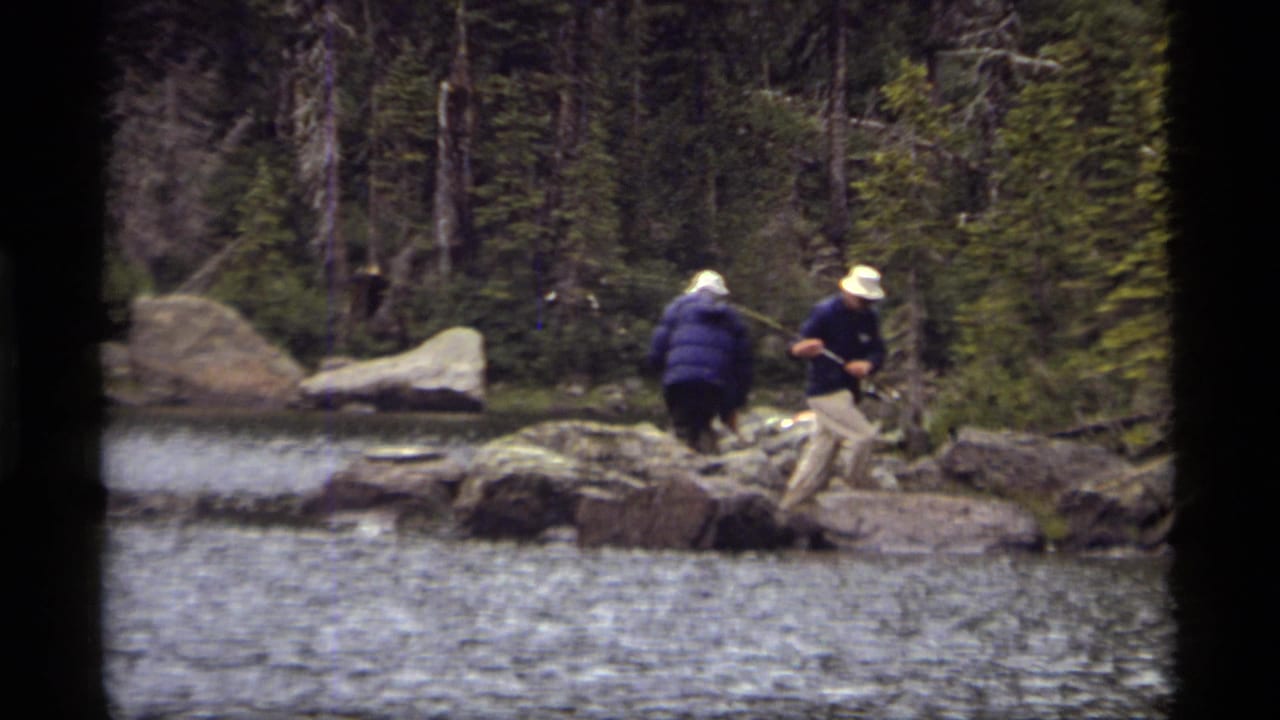 Two hat-wearing anglers move along rocks at the edge of a forested lake.