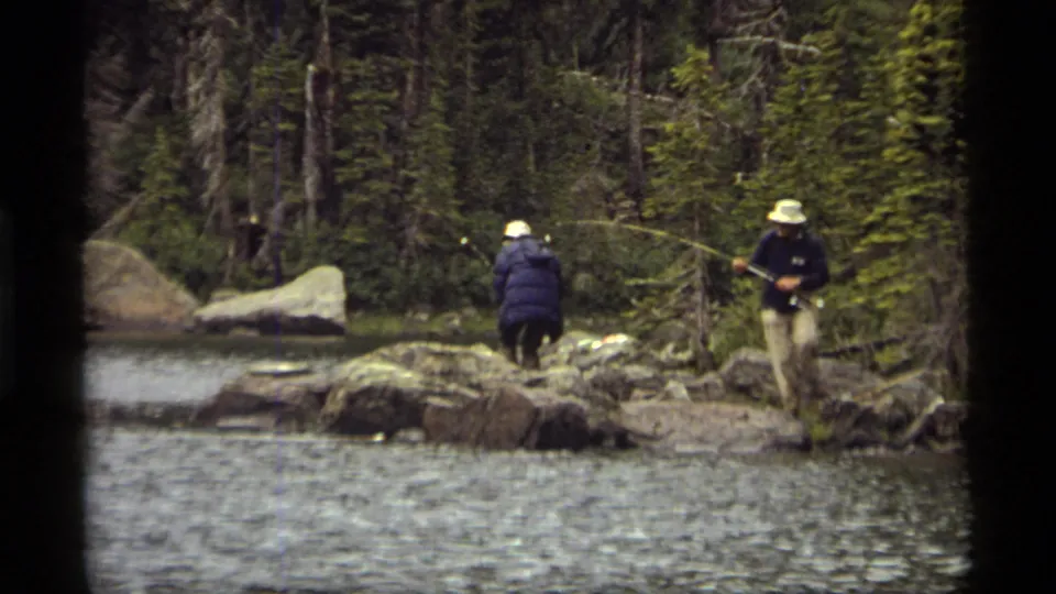 A 1977 8mm frame shows two people fishing from Sapphire Lake's rocky shoreline, with one person nearer the water and forest behind.