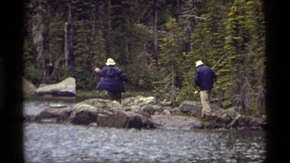 Two people fish from separate spots along Sapphire Lake's rocky Montana shore, surrounded by water, boulders, and evergreen trees.