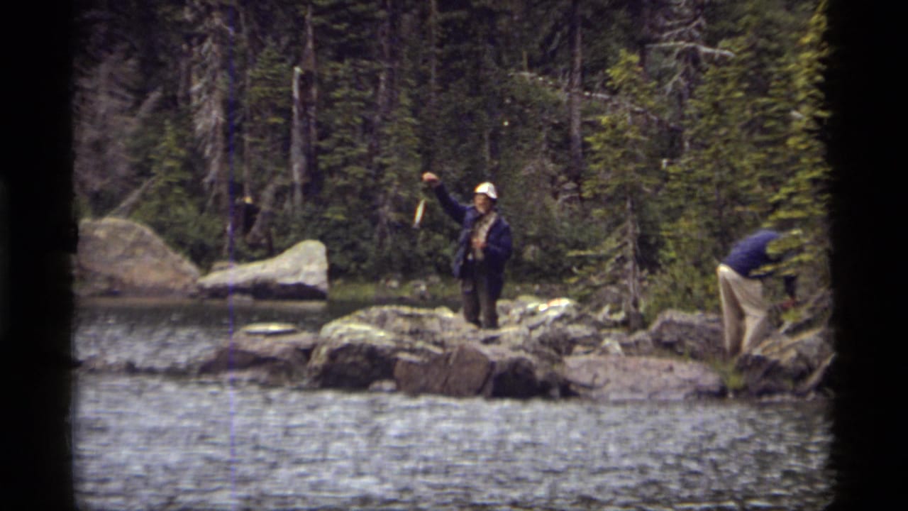 1977 Montana home-movie frame showing men on a rocky Sapphire Lake shore, with one holding up a small fish near the water.