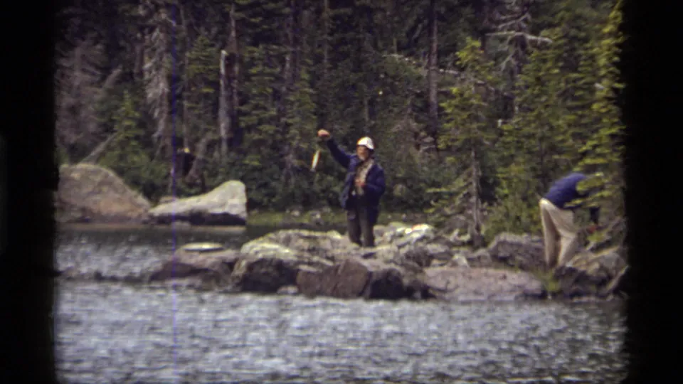1977 Montana home-movie frame showing men on a rocky Sapphire Lake shore, with one holding up a small fish near the water.
