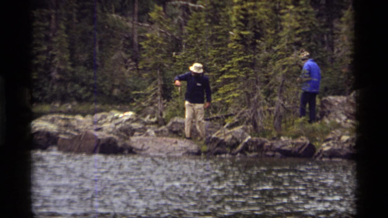 Two men stand on a rocky bank near water with evergreen trees behind them.