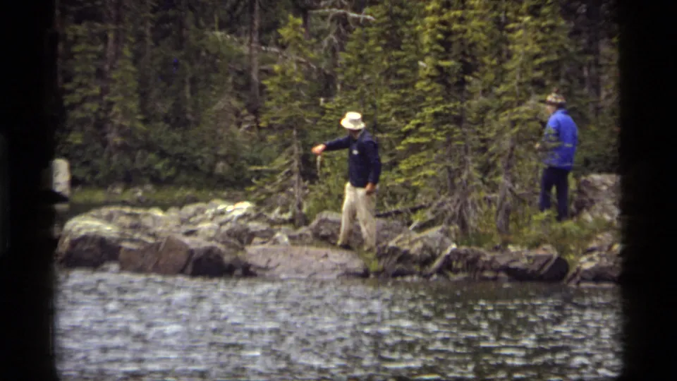 A 1977 8mm frame shows two men on the Sapphire Lake shoreline, one gesturing toward the water while another stands near trees.