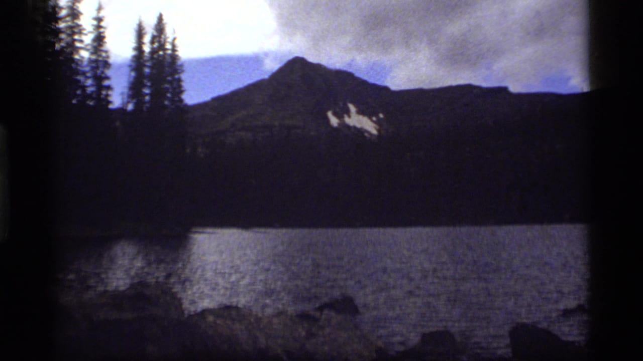 1977 8mm view of Sapphire Lake, Montana, with still water, dark tree-covered slopes, snow patches, and a cloudy mountain sky.