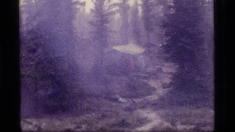 1977 Sapphire Lake home-movie frame showing a tent beside a damp path in a wooded, rain-softened campsite.