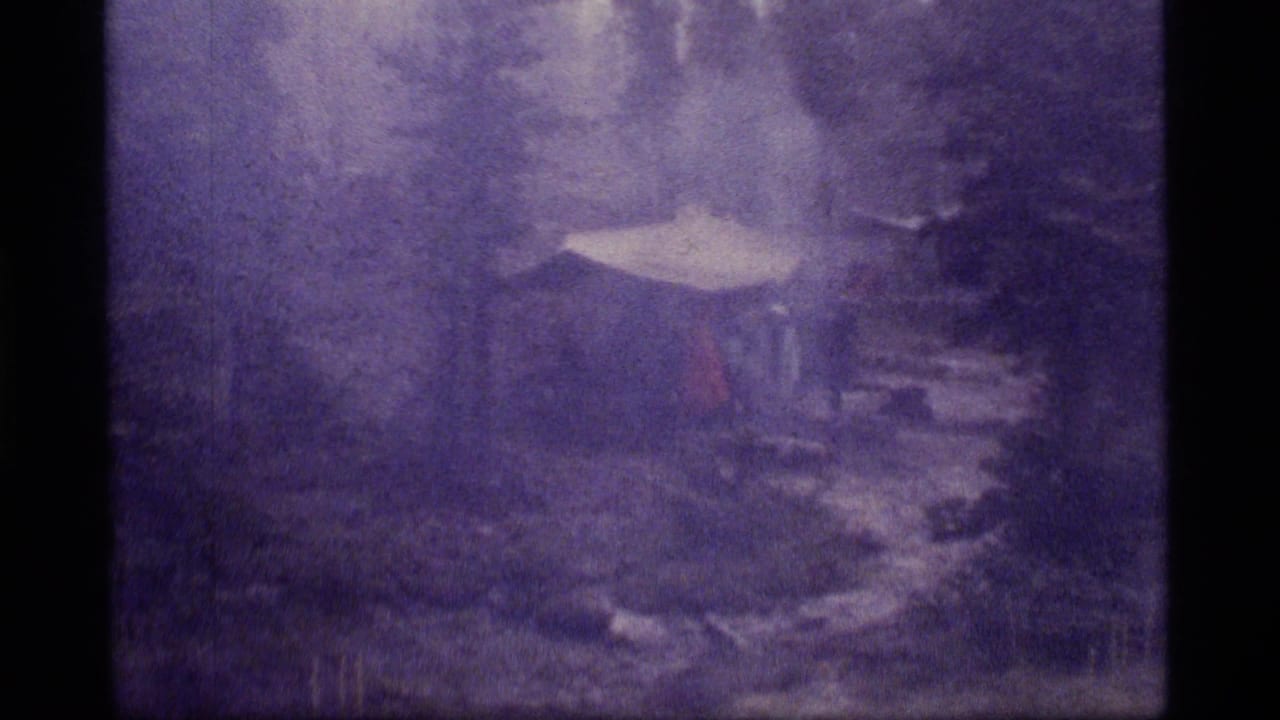 1977 8mm frame from Sapphire Lake, Montana, showing a snowy wooded campsite with a pale shelter shape and stormy haze.