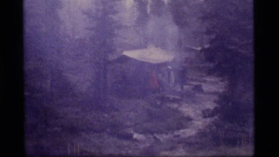 1977 8mm frame from Sapphire Lake, Montana, showing a snowy wooded campsite with a pale shelter shape and stormy haze.