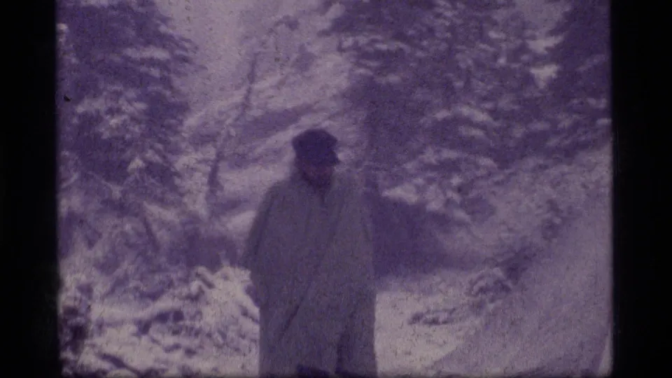 The adult appears near the camera in a snowy evergreen setting, with the face indistinct in the 8mm frame.