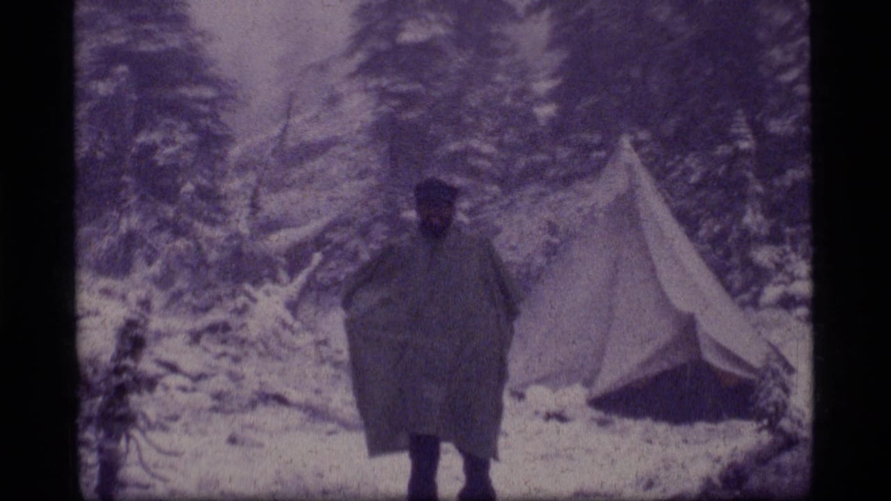 Lone camper in dark poncho walking near a tent surrounded by snow and trees.