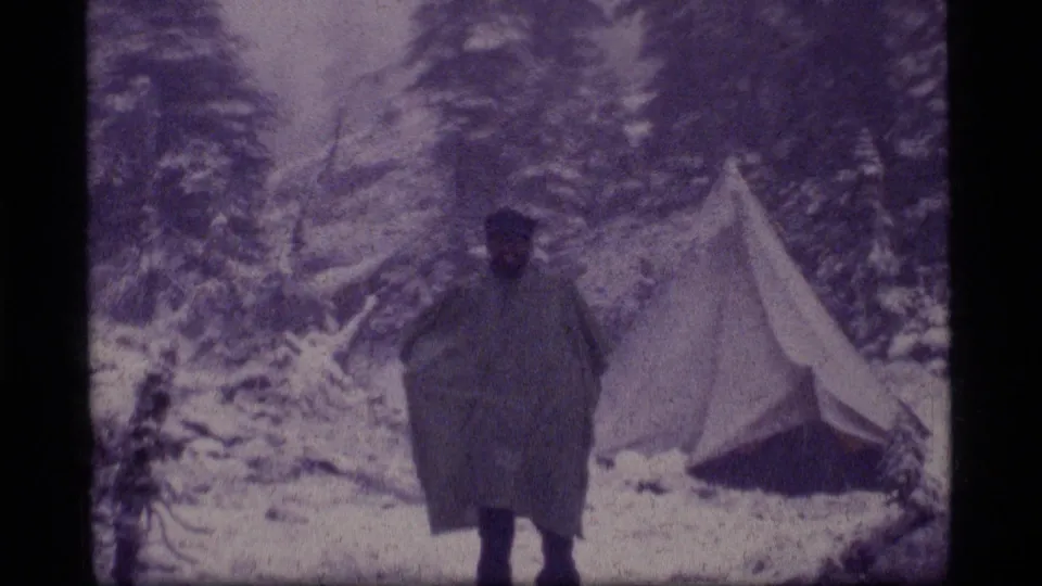 1977 Sapphire Lake, Montana frame of a camper moving through snow near a tent in a forested mountain setting.