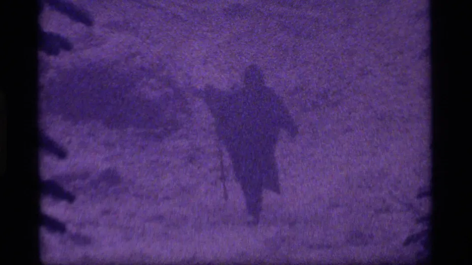 Purple-tinted 1977 8mm frame from Sapphire Lake, Montana, showing a hooded figure in snow near evergreen branches with a shovel-like tool.