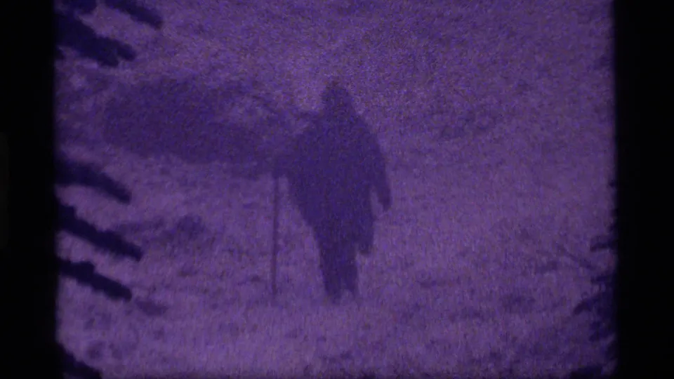 In 1977 Sapphire Lake, Montana footage, a parka-clad figure appears in deep snow amid dark evergreen branches and purple-shifted 8mm grain.