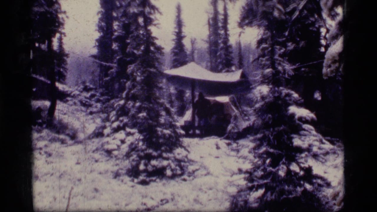1977 8mm frame from Sapphire Lake, Montana showing a snow-covered forest campsite and tarp shelter among pines.