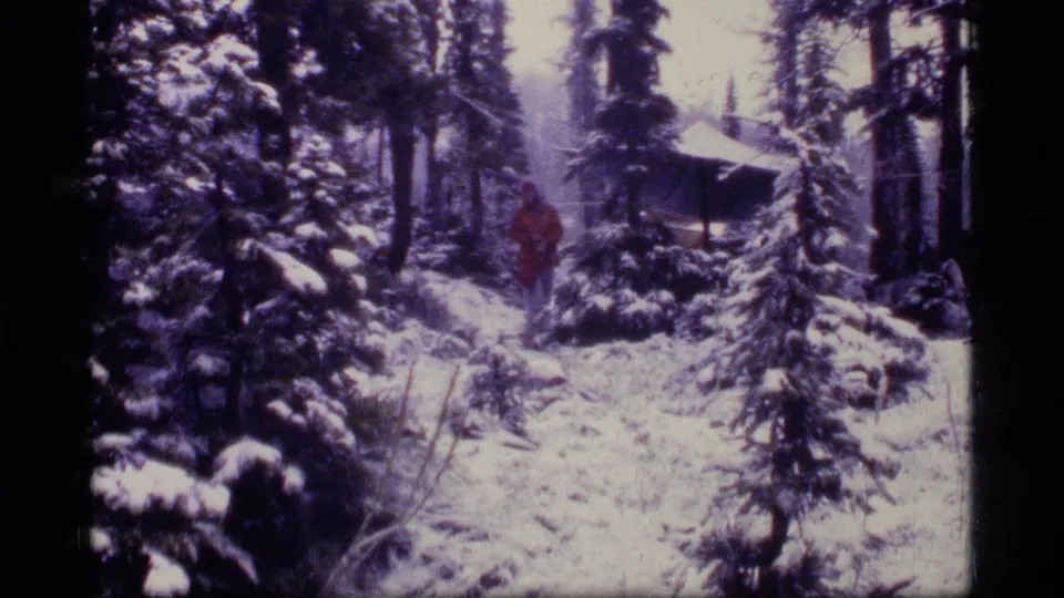 A man in a red jacket walks through the snowy Sapphire Lake campsite near a tarp shelter and pine trees.