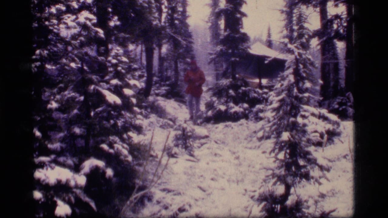 Man in a red coat walking through snow-covered evergreen woods with a small cabin behind him.