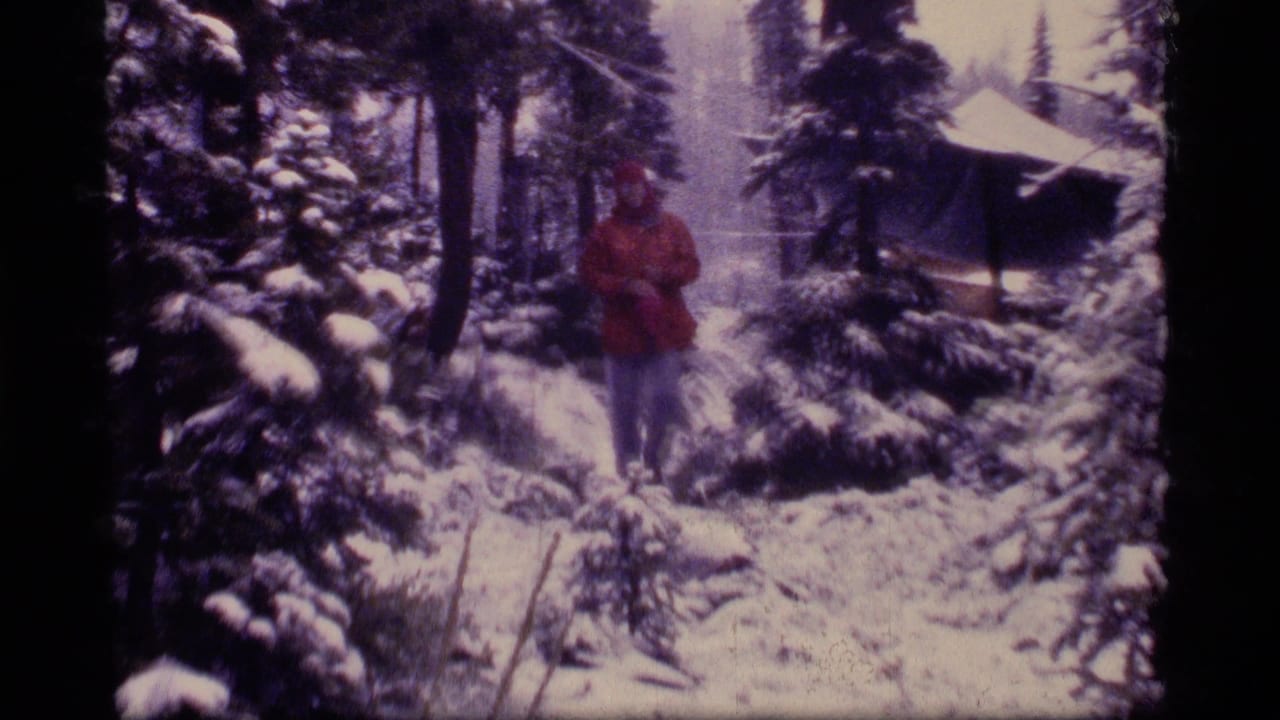 1977 Montana frame with a red-coated man walking through deep snow in evergreen woods, with a cabin roof visible at right.