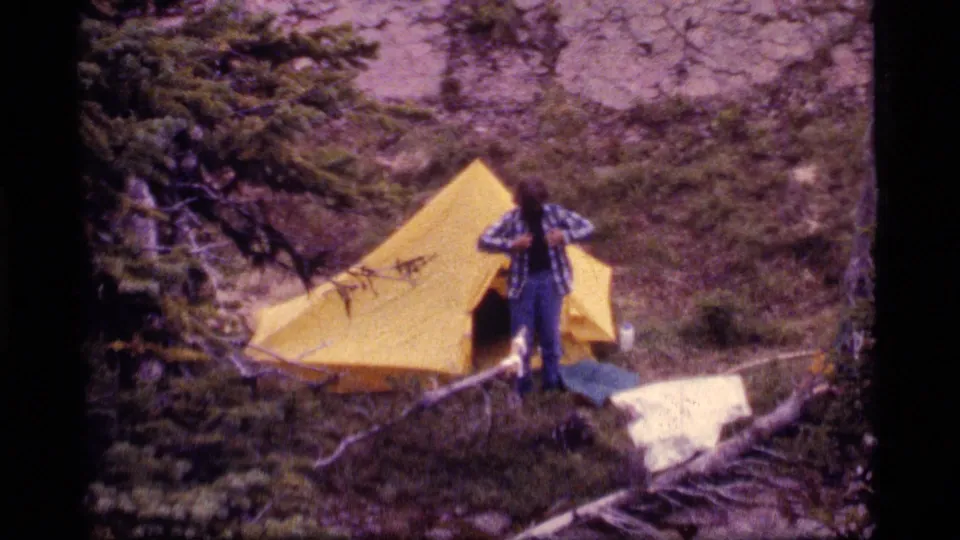 1977 home-movie frame shows a camper in plaid and jeans adjusting clothing beside a yellow tent at Sapphire Lake, Montana.