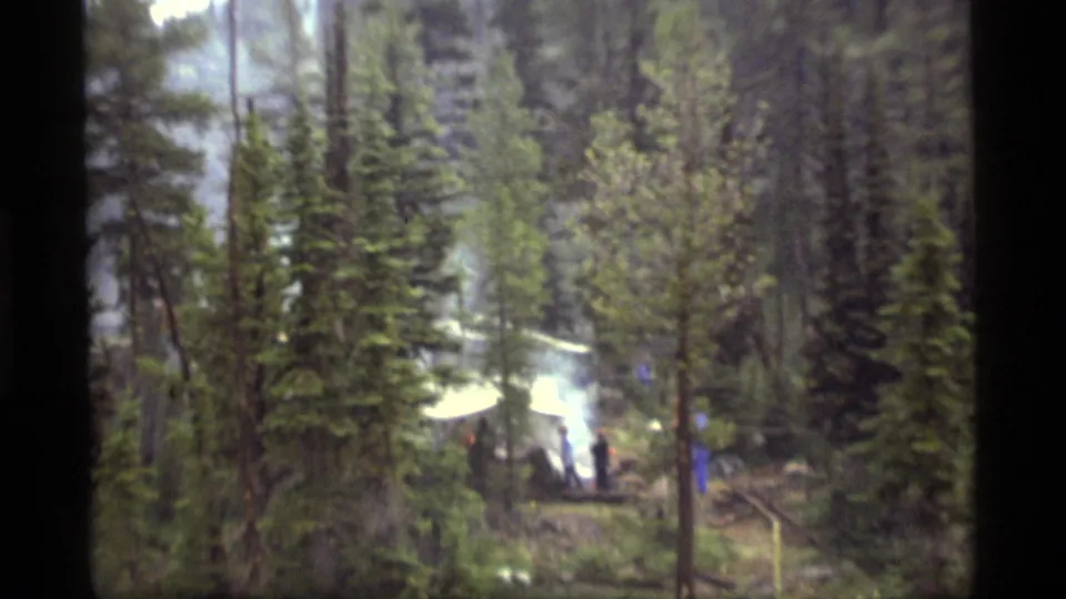 Wide 8mm view of the Sapphire Lake forest campsite, with distant people, smoke, and a white canopy among tall pines.