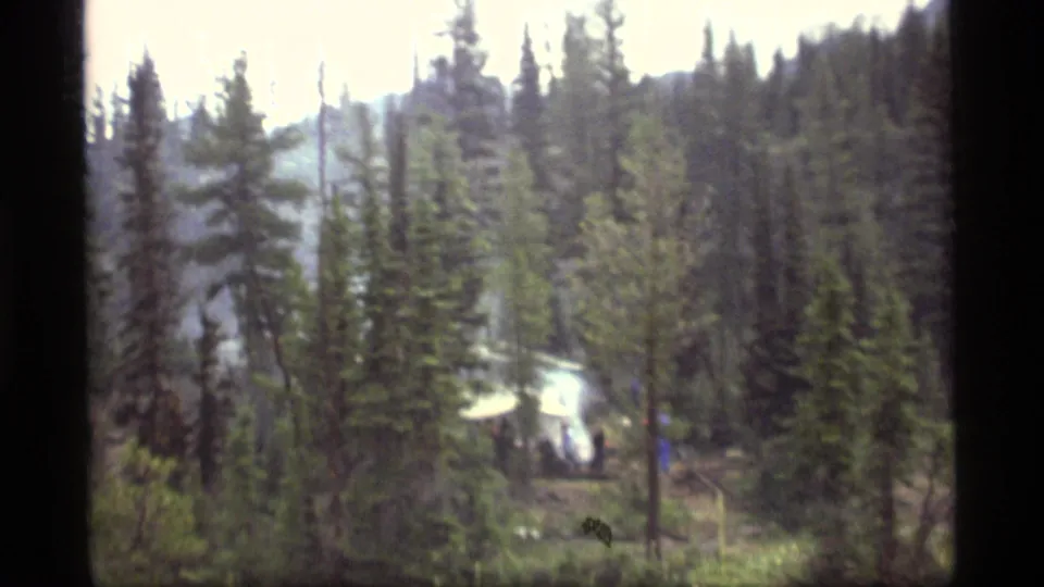 1977 Sapphire Lake campsite seen through dense Montana pines, with a white canopy, faint people, and smoke in daylight.