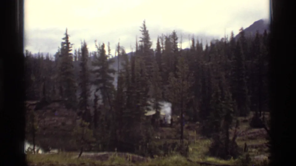 1977 home-movie frame of a shaded Montana forest campsite, with smoke and a small structure partly obscured by trees.