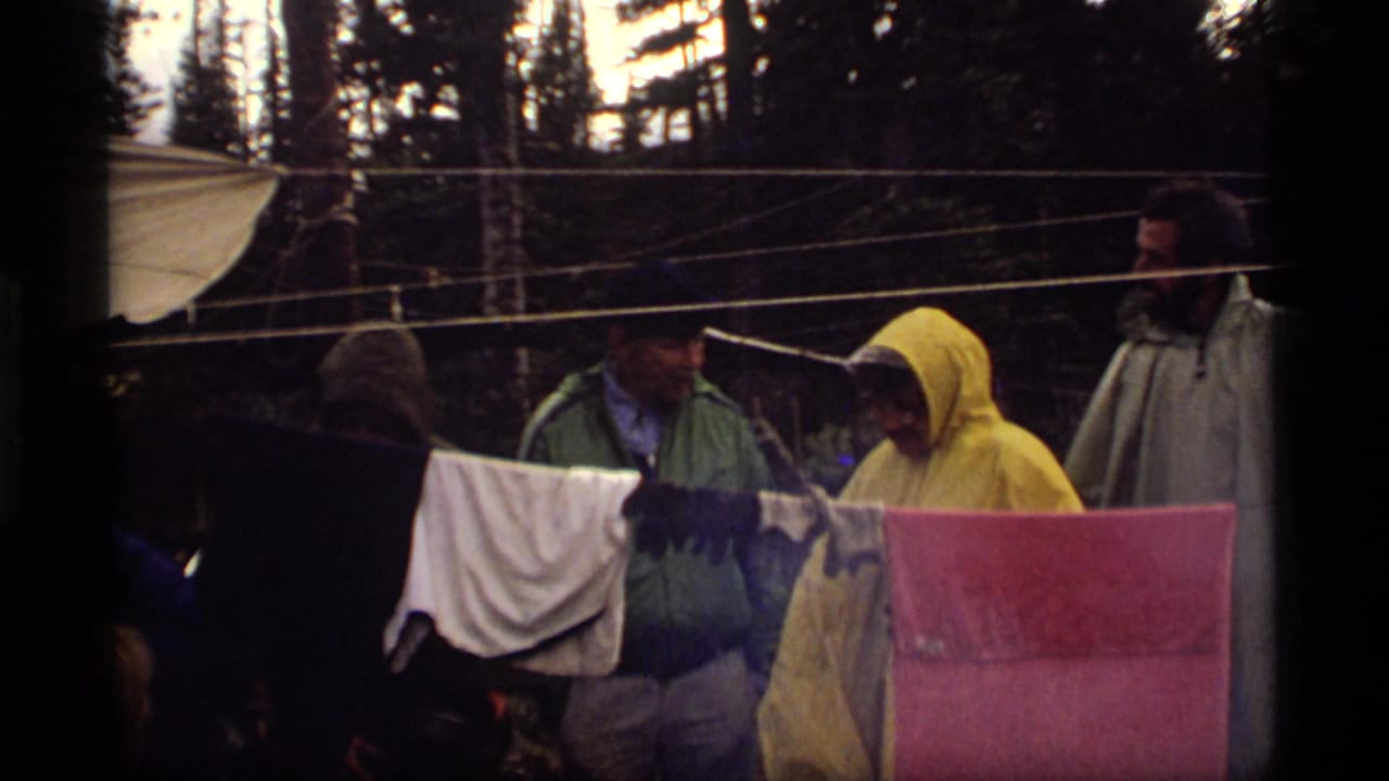 Three campers stand by a clothesline with damp garments in a wooded campsite.
