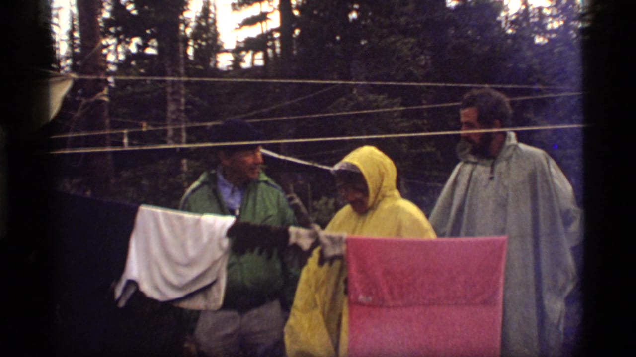 Three campers at the 1977 Sapphire Lake campsite stand behind a clothesline, with a yellow poncho and gray poncho prominent in the frame.