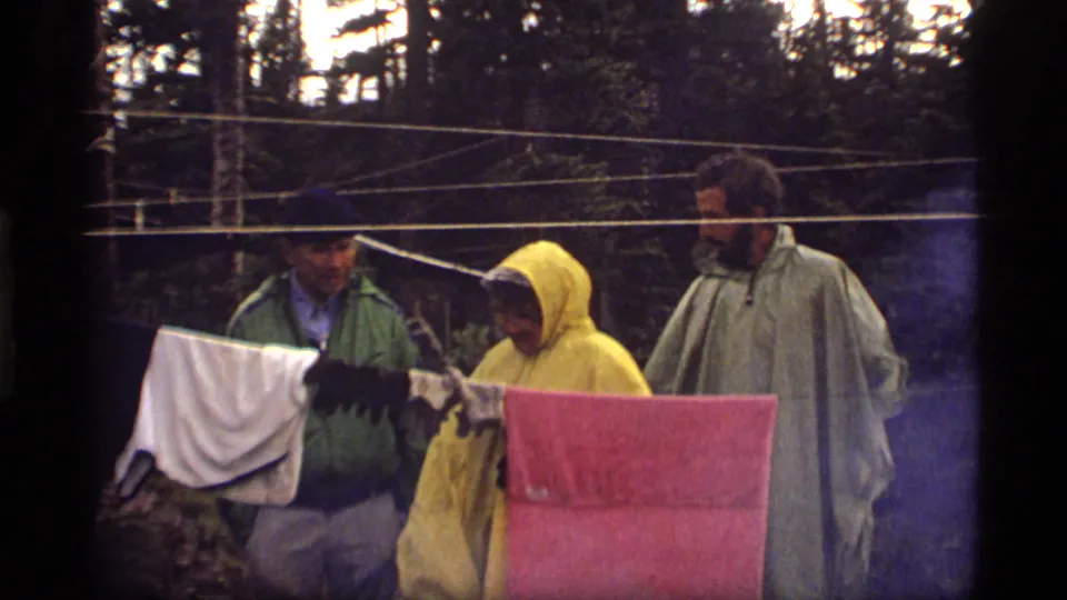 In the 1977 Montana home movie, campers stand behind a campsite clothesline while damp white and pink fabric hangs in the foreground.