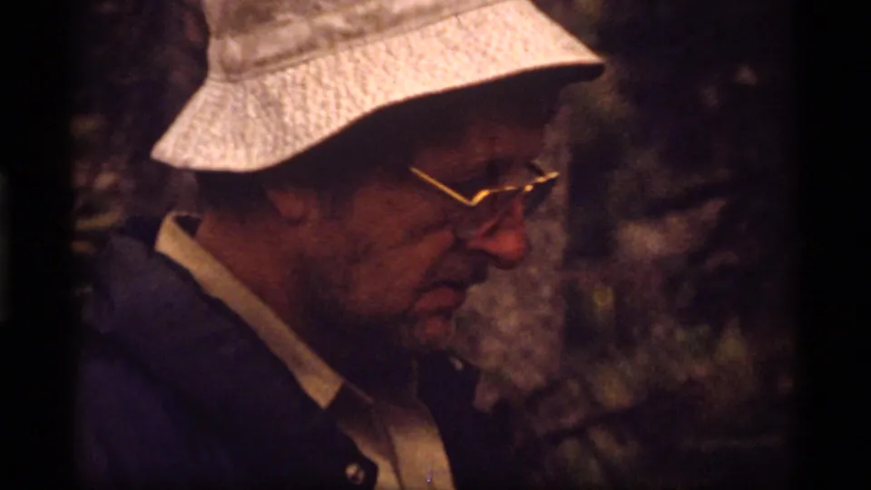 Close-up from the 1977 Sapphire Lake footage shows a man in glasses and a light cap, framed in soft, dark 8mm forest light.