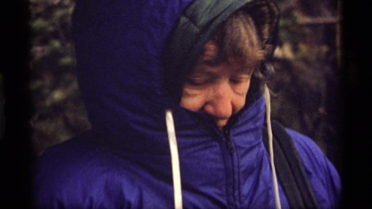 Older person in a bright blue hooded puffy jacket stands outdoors at Sapphire Lake, Montana in 1977 home-movie footage.