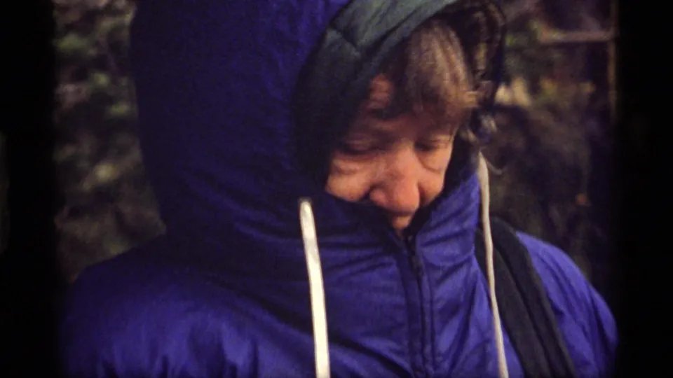 Older person in a bright blue hooded puffy jacket stands outdoors at Sapphire Lake, Montana in 1977 home-movie footage.