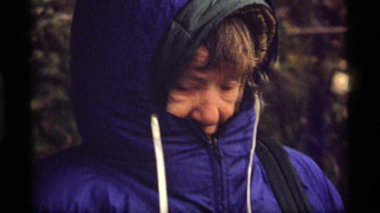 Older person in a blue hooded puffy jacket, face partly obscured by the high collar, appears in 1977 Montana 8mm footage.
