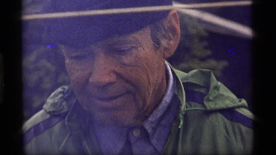 Older man wearing a cap and green jacket smiles faintly in a soft, flare-affected 1977 Montana home-movie frame.