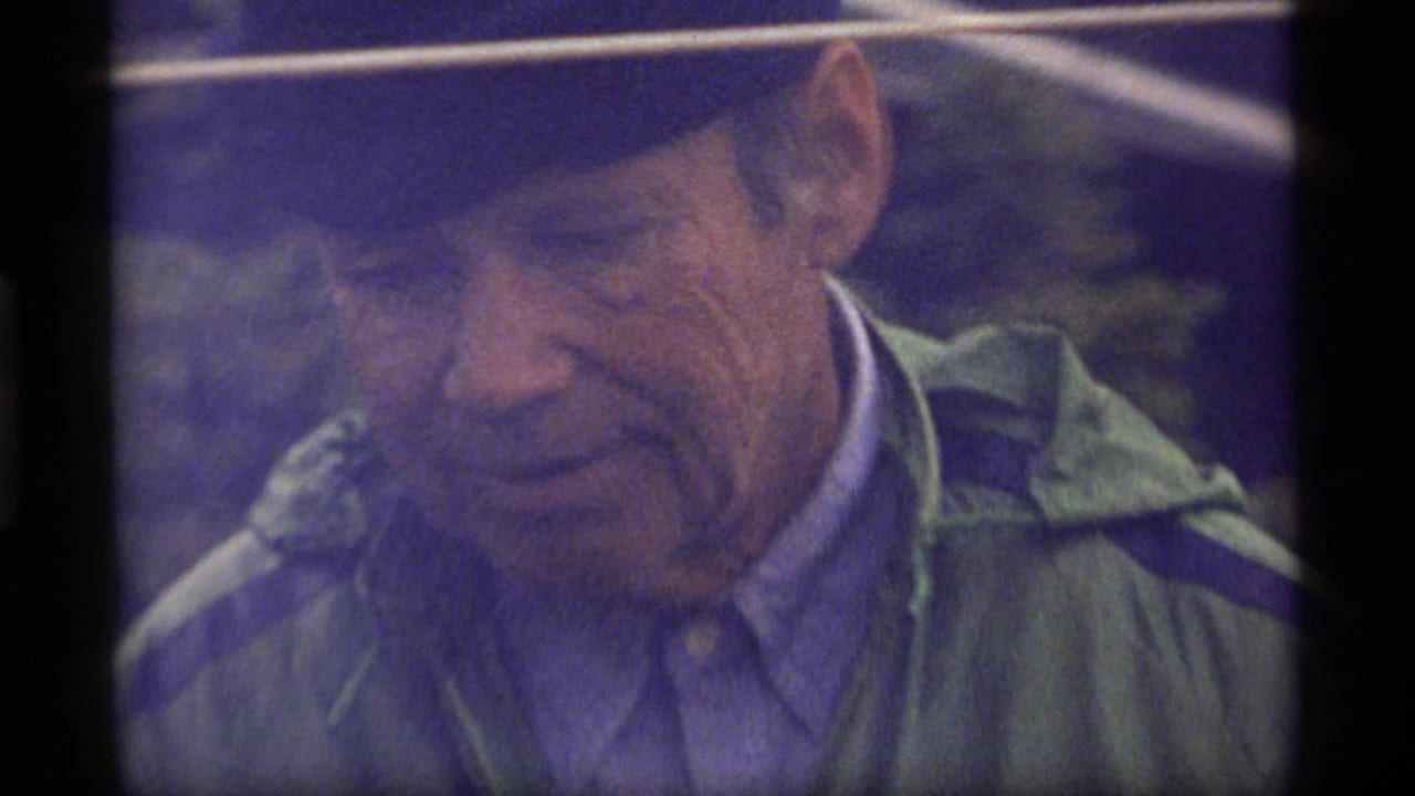 In 1977 Sapphire Lake home-movie footage, an older man in a dark hat and green jacket looks downward in a close, grainy frame.