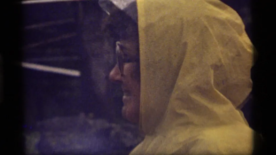 1977 Sapphire Lake 8mm frame of a woman in a yellow hooded coat and glasses, shown in side profile with dim outdoor lighting.