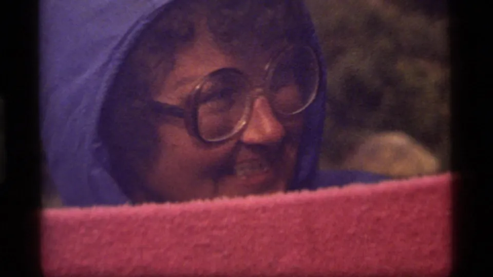 Close 1977 Sapphire Lake 8mm frame of a hooded woman with large glasses smiling behind a pink foreground barrier.