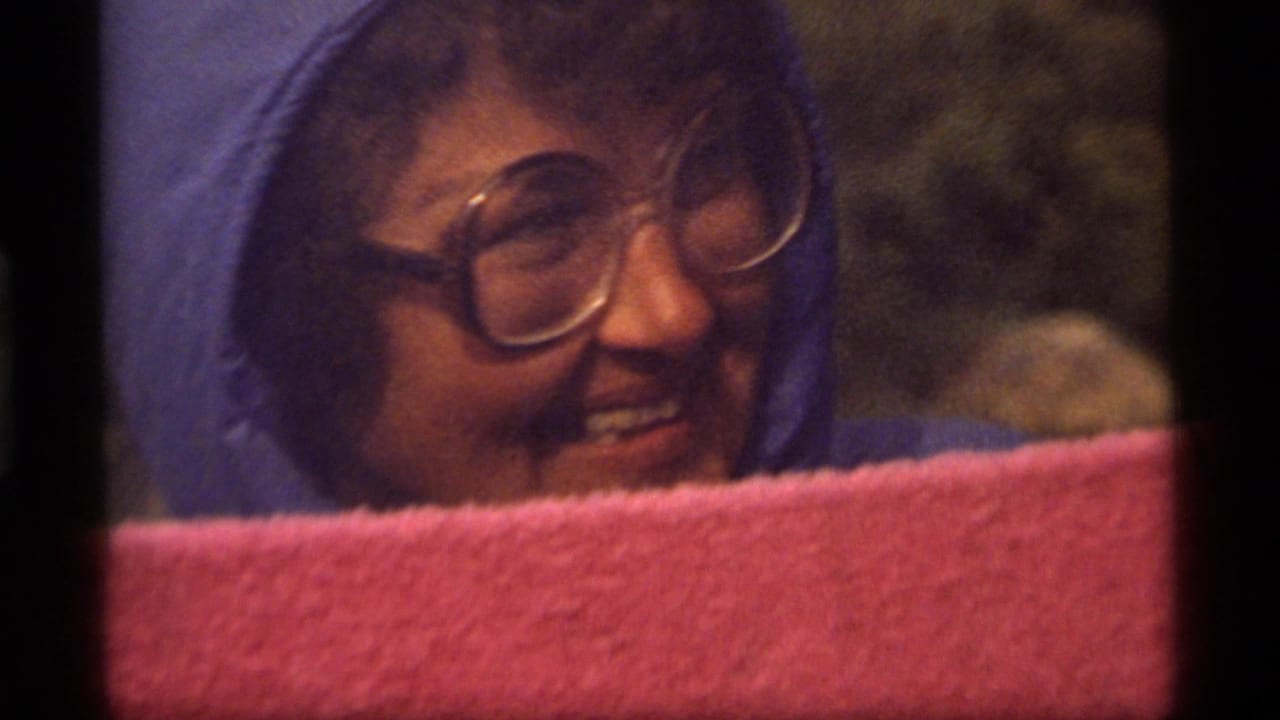 1977 Sapphire Lake home-movie frame showing a woman in a blue hooded jacket and round glasses smiling outdoors.