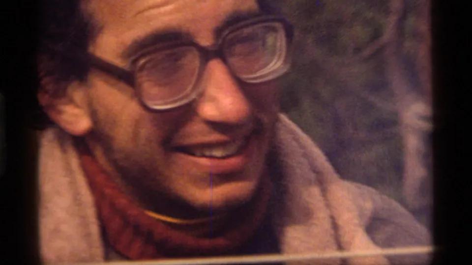 1977 Sapphire Lake home-movie frame showing a man in thick glasses, beige coat, and red turtleneck smiling outdoors.