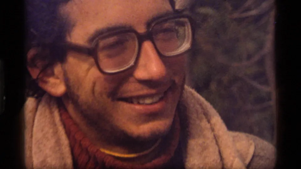 1977 Sapphire Lake home-movie frame showing a man with thick-rimmed glasses laughing in a casual outdoor setting.